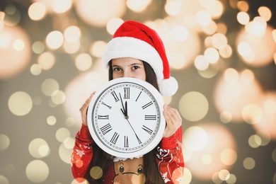 Image of New Year countdown. Cute little girl in Santa hat holding clock against blurred lights on background