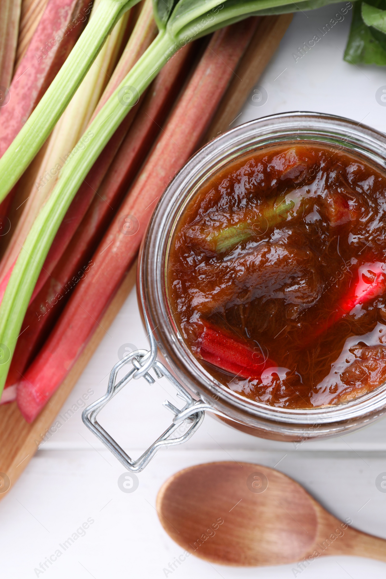 Photo of Jar of tasty rhubarb jam, stems and spoon on white wooden table, flat lay