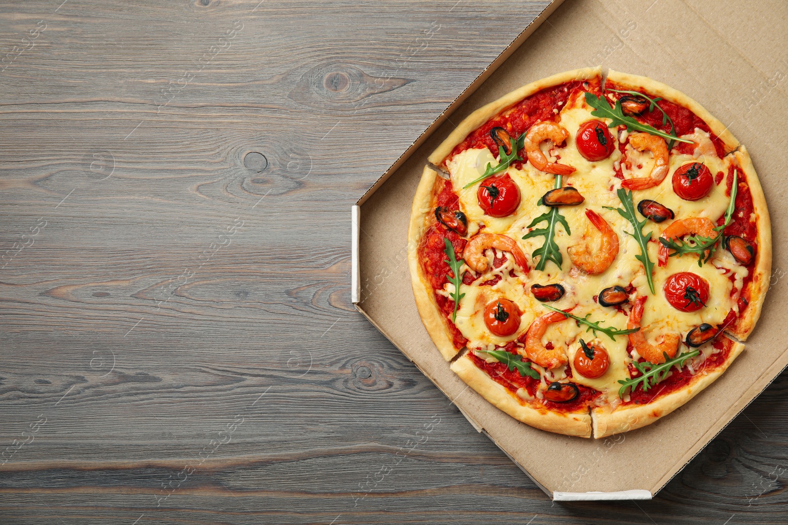 Photo of Delicious seafood pizza in cardboard box on wooden table, top view. Space for text