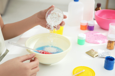 Little girl adding sparkles into homemade slime toy at table, closeup of hands