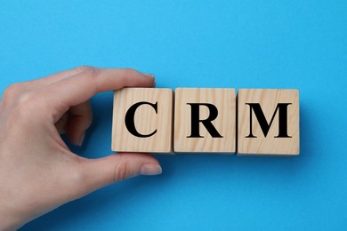 Woman making abbreviation CRM of wooden cubes on blue background, closeup. Customer Relationship Management