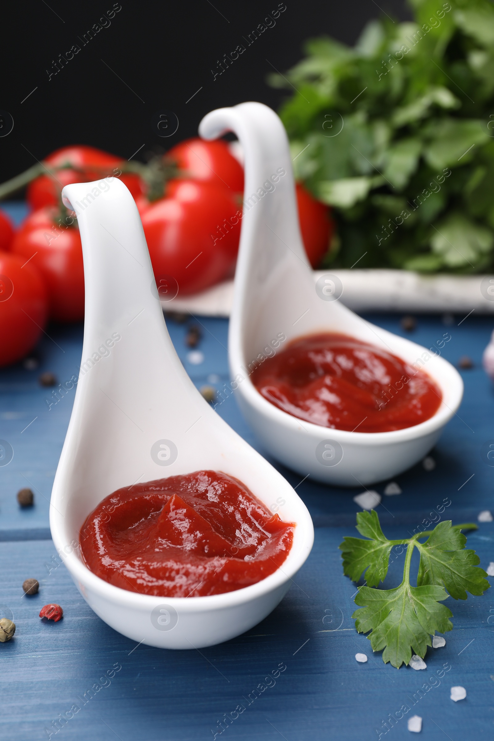 Photo of Organic ketchup in spoons and spices on blue wooden table, closeup. Tomato sauce