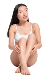 Beautiful young Asian woman applying body cream onto legs on white background