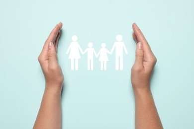 Image of Woman holding hands around paper silhouette of family on light blue background, top view. Insurance concept