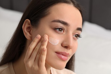 Photo of Young woman with dry skin applying cream onto her face on blurred background