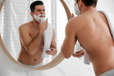 Handsome man applying shaving foam near mirror in bathroom