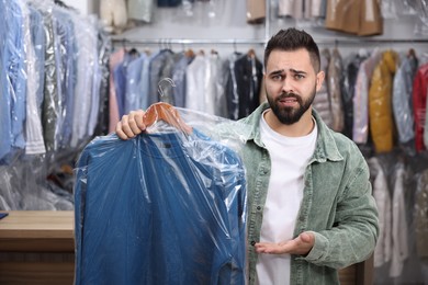 Dry-cleaning service. Displeased man holding hanger with sweatshirt in plastic bag indoors