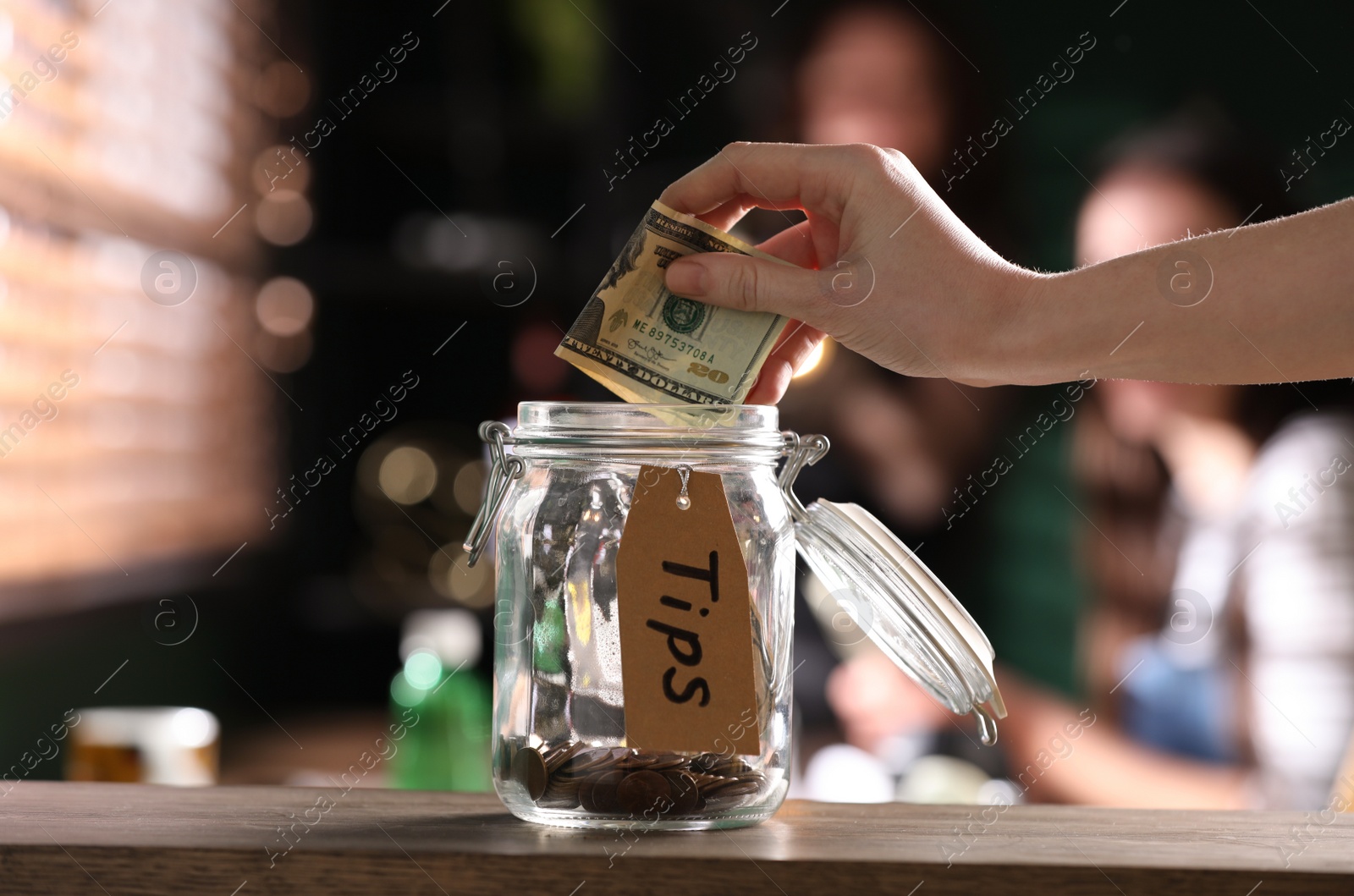 Photo of Woman putting tips into glass jar on wooden table indoors, closeup