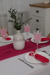 Photo of Table setting. Glasses of tasty beverage, plates, pink napkins and vase with green branches in dining room