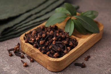 Wooden tray with aromatic cloves and green leaves on brown table, closeup