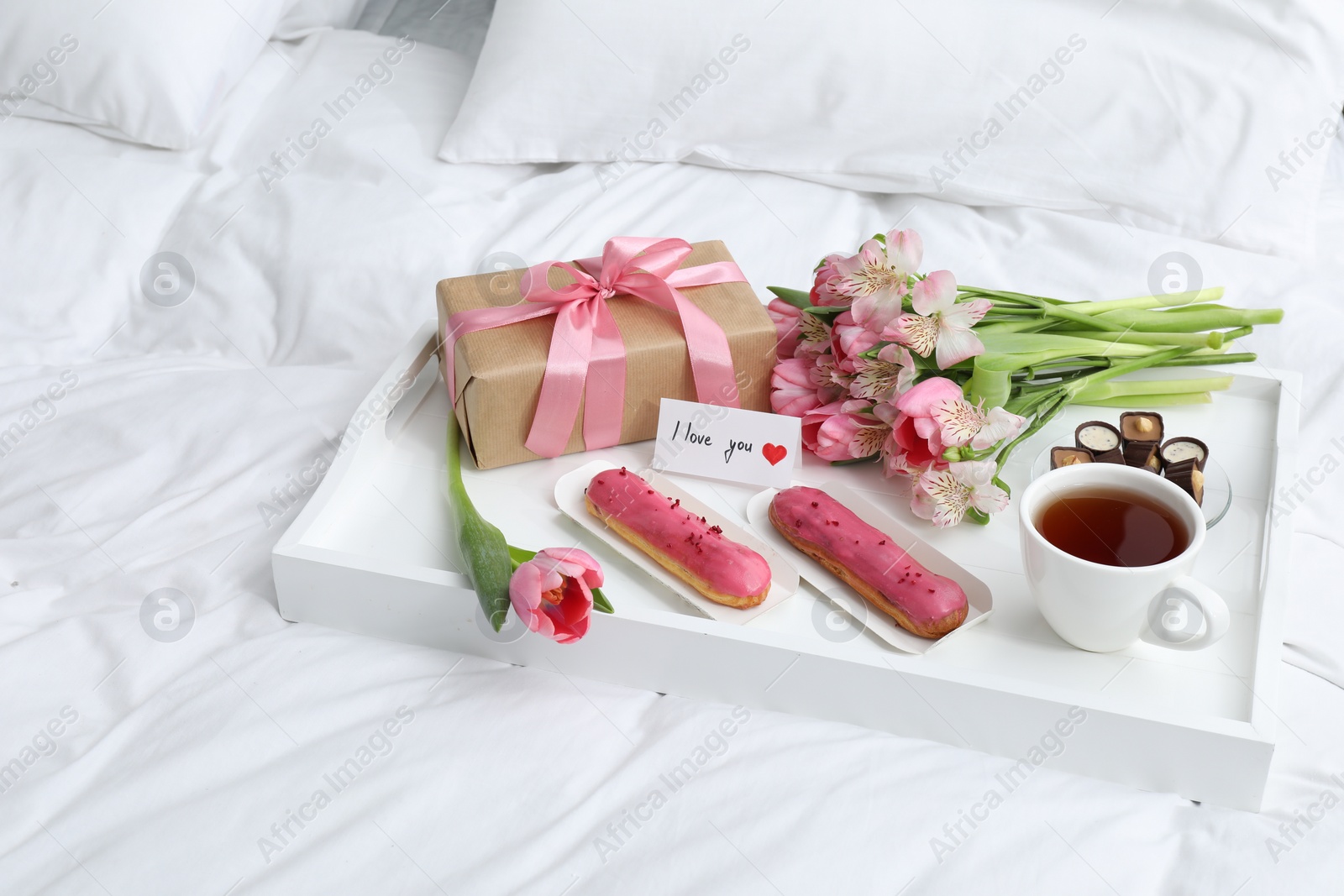 Photo of Tasty breakfast served in bed. Delicious desserts, tea, flowers, gift box and card with phrase I Love You on tray