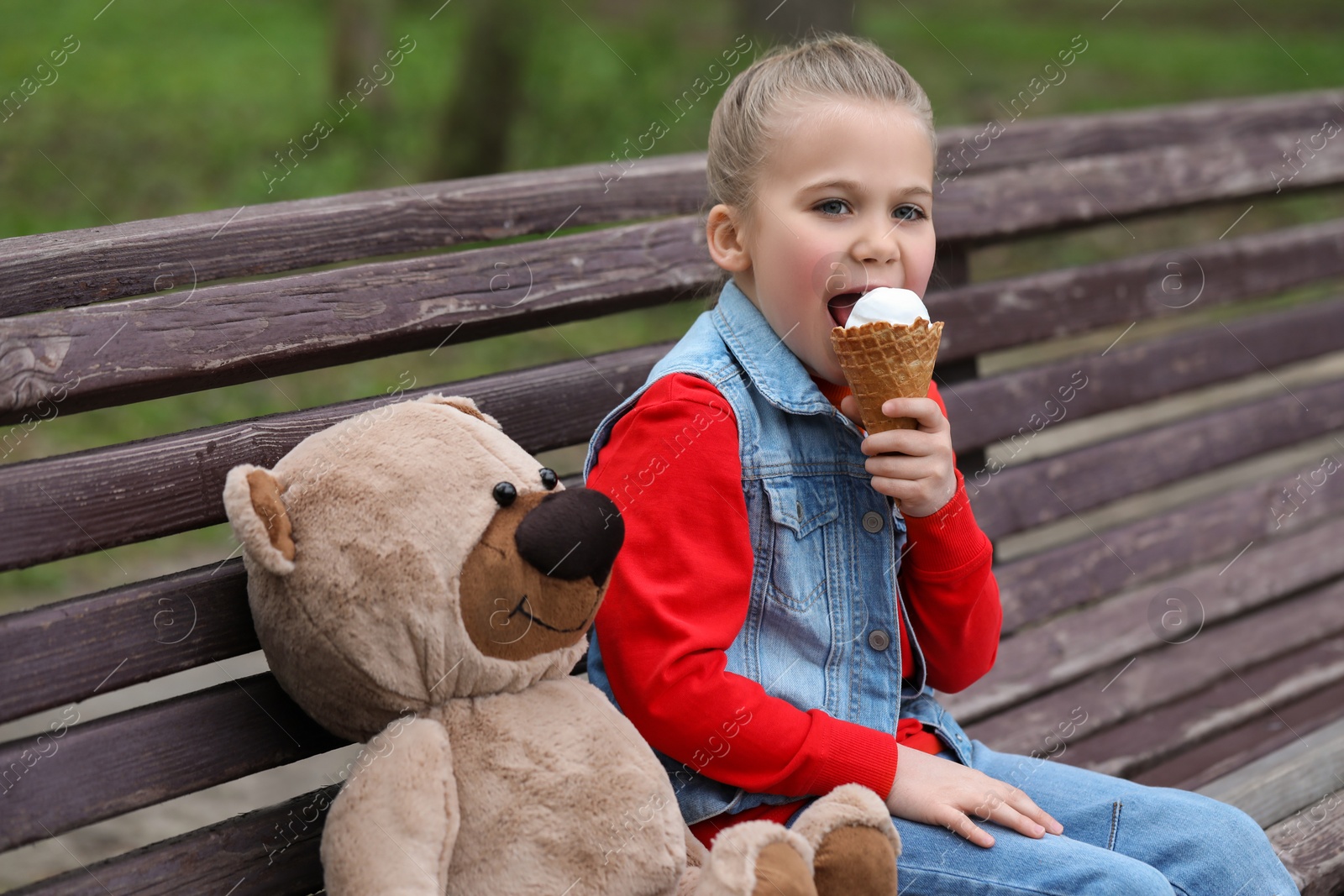 Photo of Little girl with teddy bear eating ice cream on wooden bench outdoors