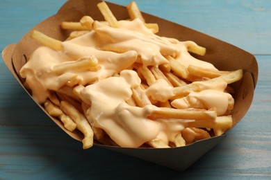 Tasty potato fries and cheese sauce in paper container on light blue wooden table, closeup