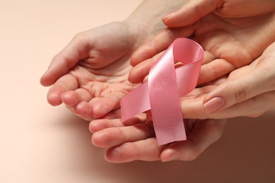 Women holding pink ribbon on beige background, closeup. Breast cancer awareness