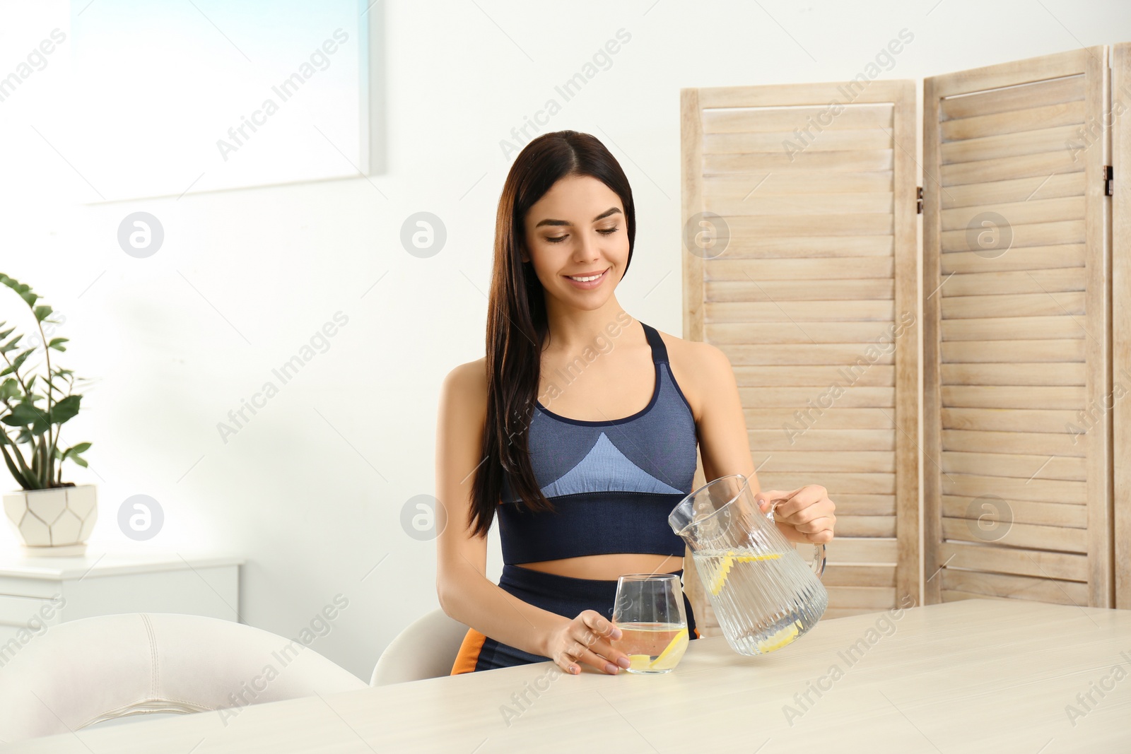 Photo of Beautiful sportive woman pouring lemon water into glass from jug at table indoors