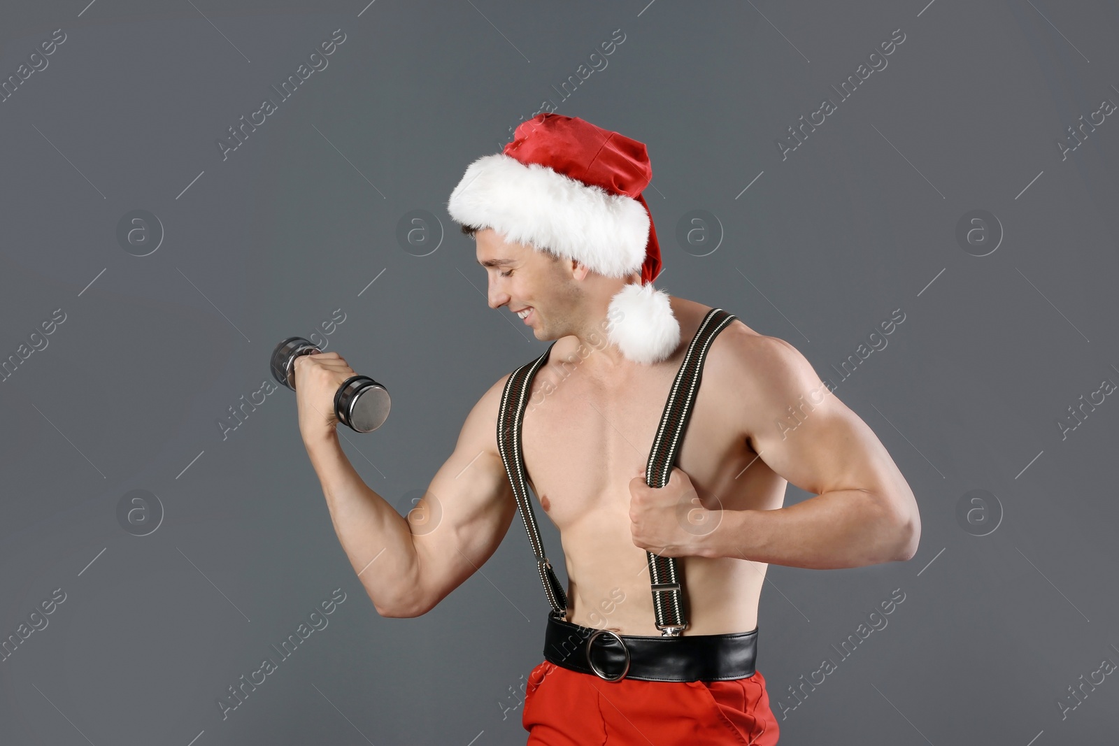 Photo of Young muscular man in Santa hat with dumbbells on gray background