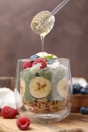 Pouring honey from dipper onto tasty chia matcha pudding with oatmeal and fruits at wooden table. Healthy breakfast