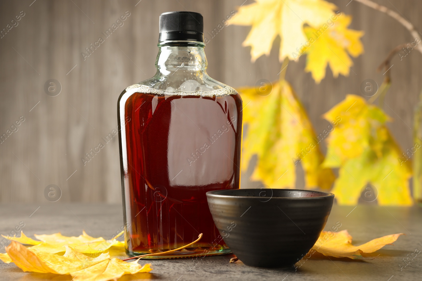 Photo of Bottle of tasty maple syrup, bowl and dry leaves on light grey table