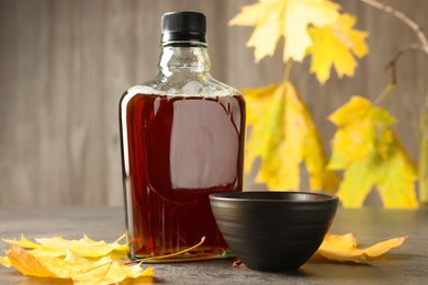 Bottle of tasty maple syrup, bowl and dry leaves on light grey table