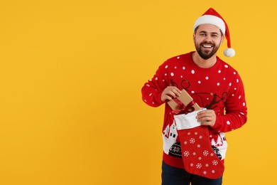 Photo of Happy young man in Christmas sweater and Santa hat taking gift from stocking on orange background. Space for text