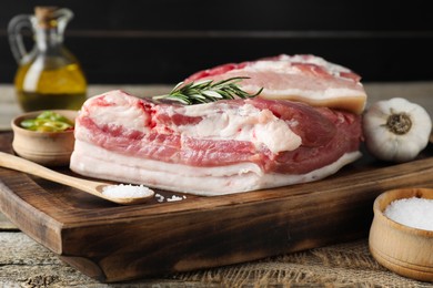 Photo of Pieces of raw pork belly, salt, oil, garlic and rosemary on wooden table, closeup