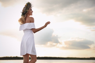 Young woman wearing wreath made of beautiful flowers near river on sunny day