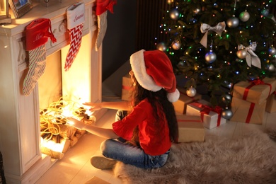 Cute little child in Santa hat sitting near Christmas tree and warming up hands in front of decorative fireplace at home