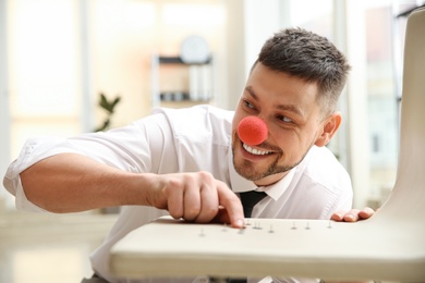 Photo of Man with clown nose putting pins on colleague's chair in office. Funny joke