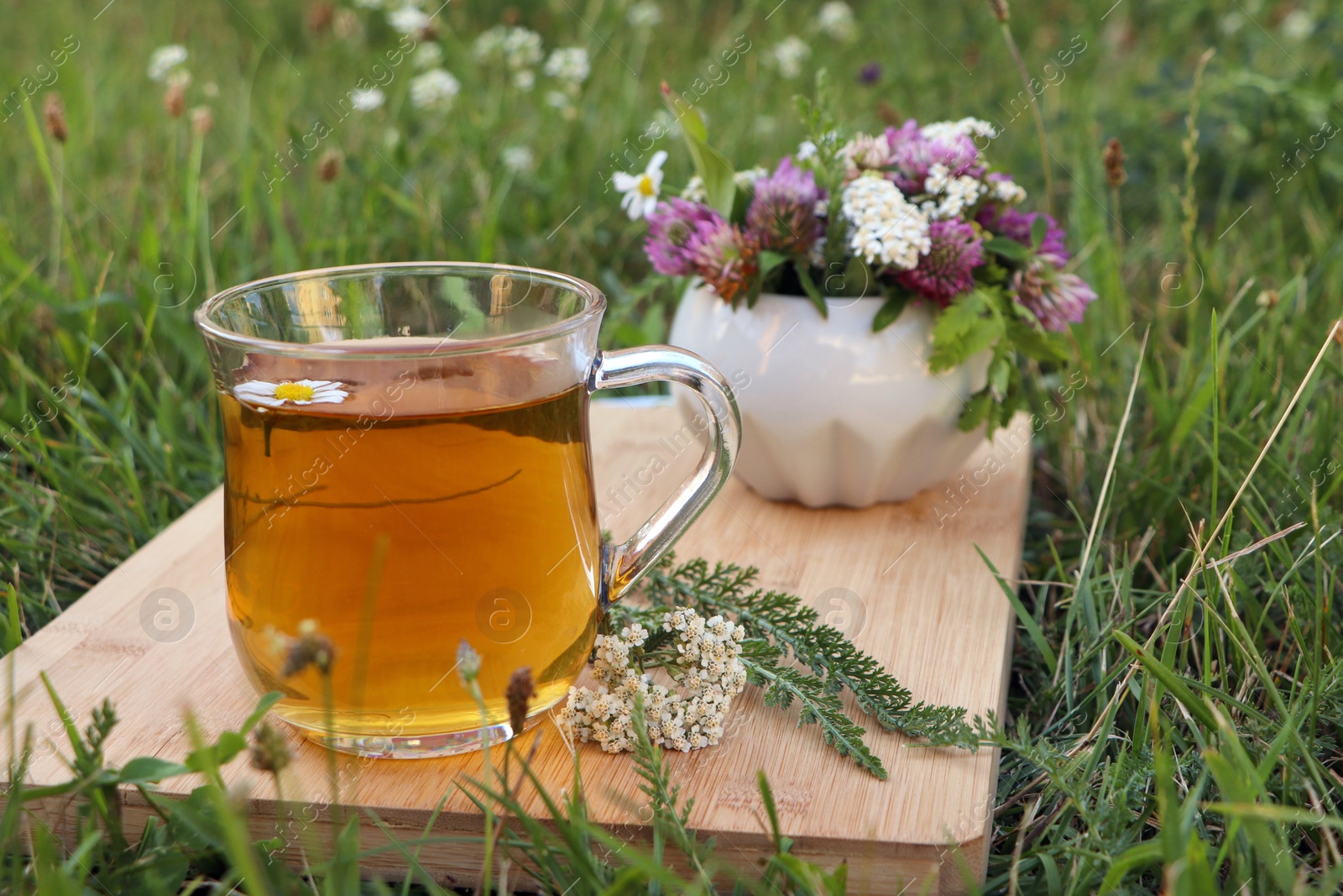 Photo of Cup of aromatic herbal tea and ceramic mortar with different wildflowers on green grass outdoors. Space for text
