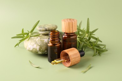 Photo of Aromatic essential oils in bottles, rosemary, pipette and pebble stones on green background