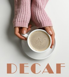 Woman holding cup of decaf coffee at white table, top view