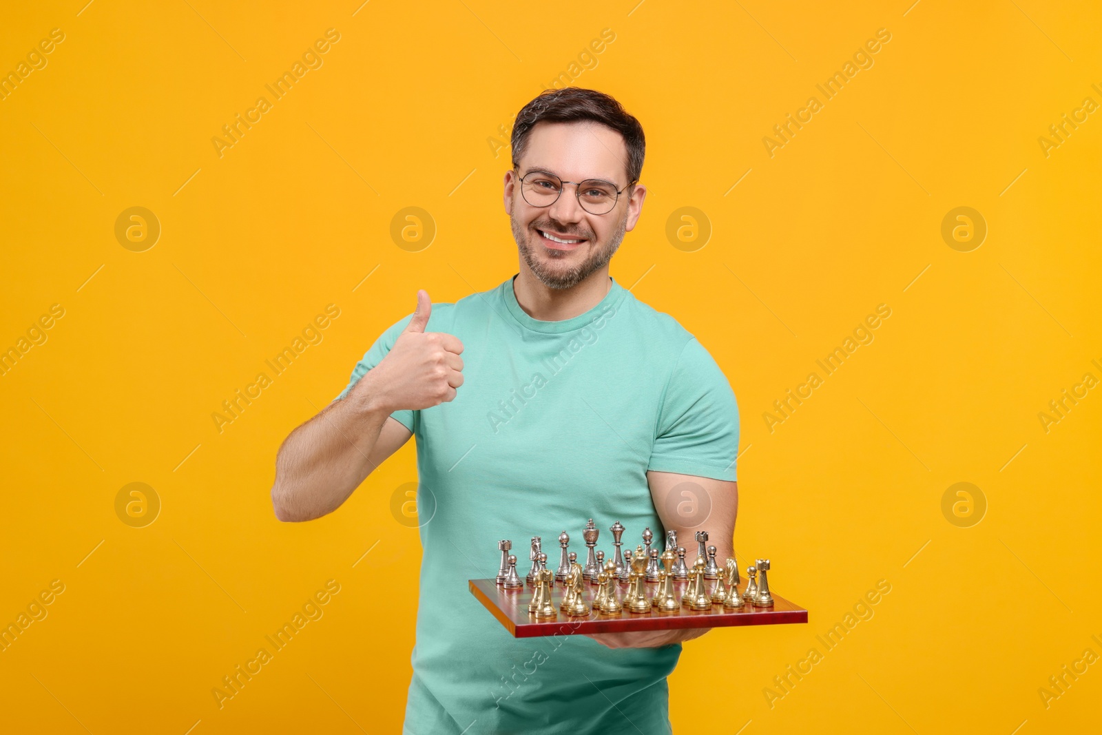 Photo of Smiling man holding chessboard with game pieces and showing thumbs up on orange background