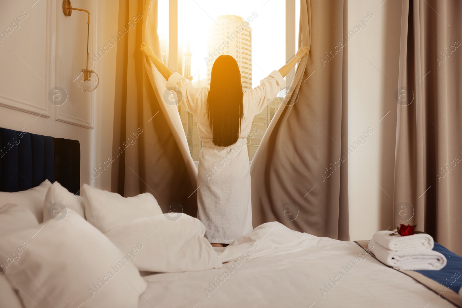 Image of Young woman opening window curtains in hotel room, back view