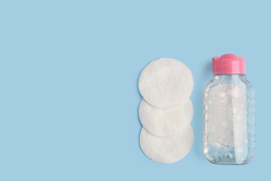 Bottle of makeup remover and cotton pads on light blue background, flat lay. Space for text