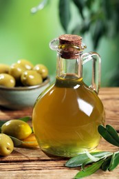 Photo of Jug of cooking oil, olives and green leaves on wooden table