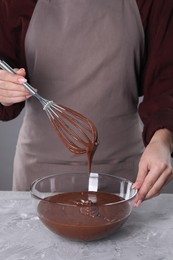 Woman with whisk mixing chocolate cream at table against grey background, closeup