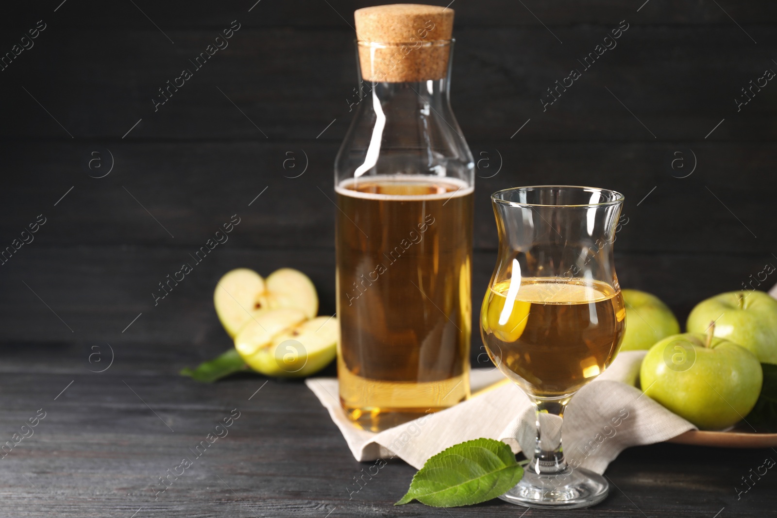 Photo of Delicious cider and apples with green leaves on black wooden table, space for text