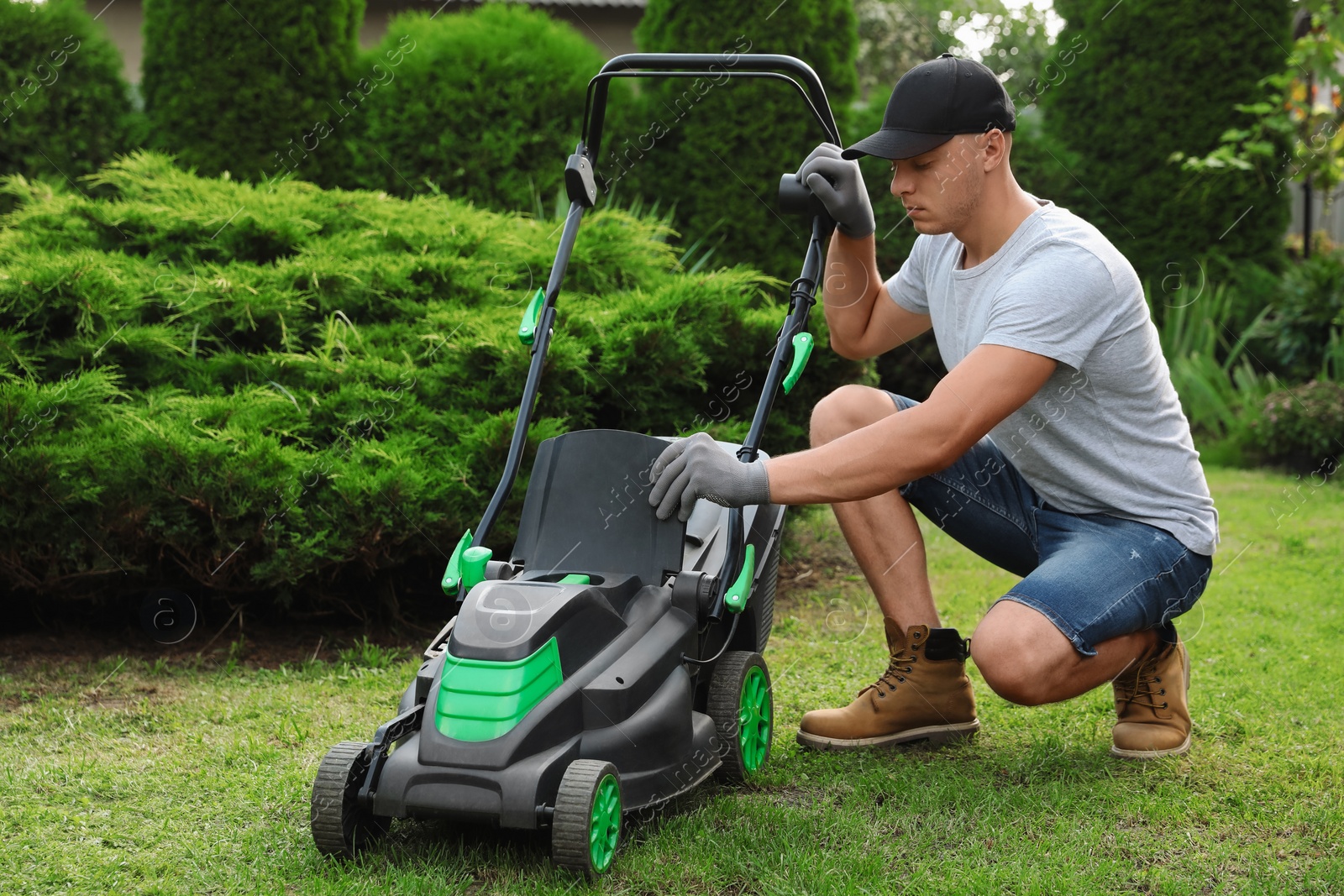 Photo of Cleaning lawn mower. Young man detaching grass catcher from device in garden