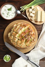 Freshly baked bread with tofu cheese, green onions, sauce and knife on wooden table, flat lay