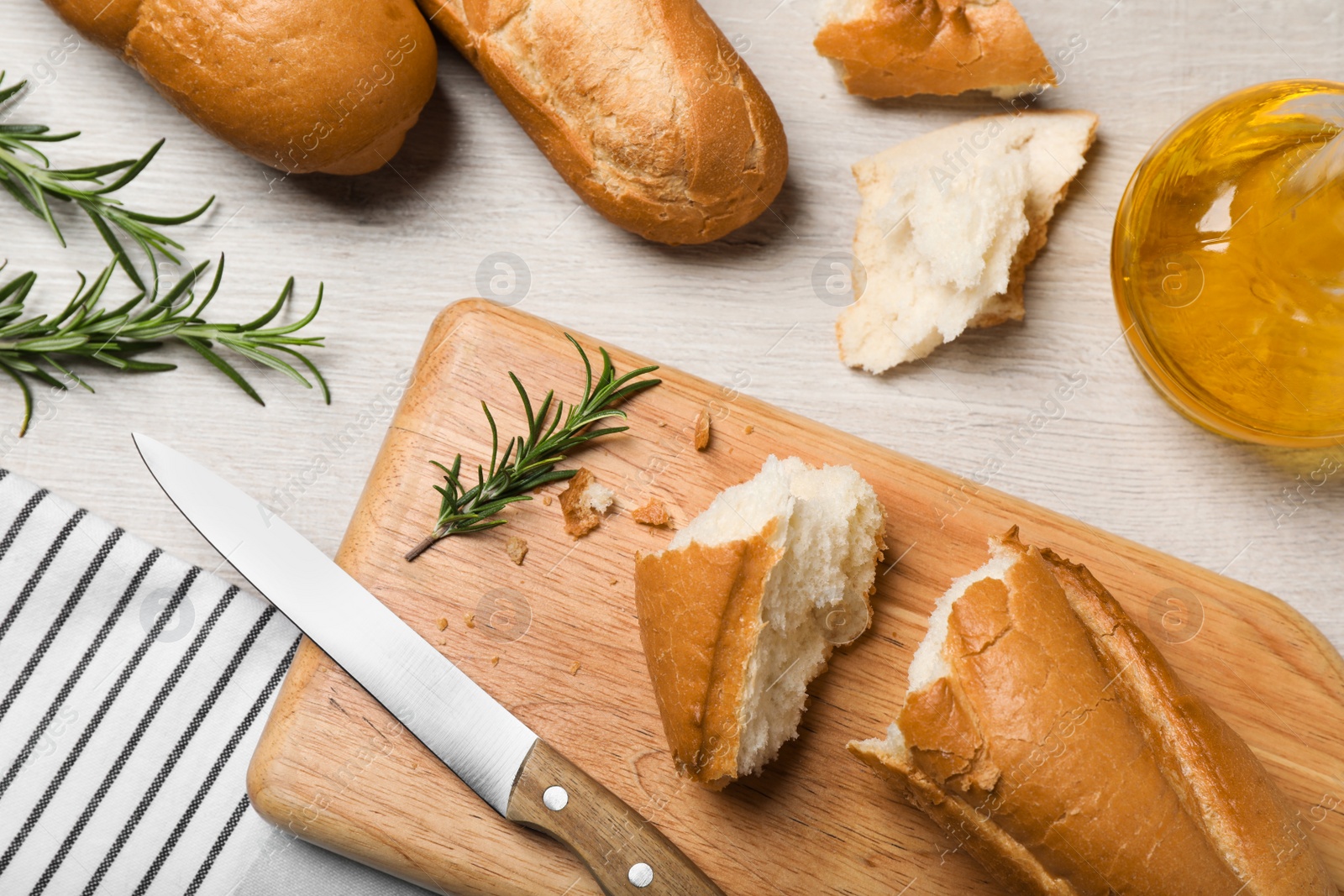 Photo of Broken tasty baguette, knife and rosemary on white wooden table, flat lay