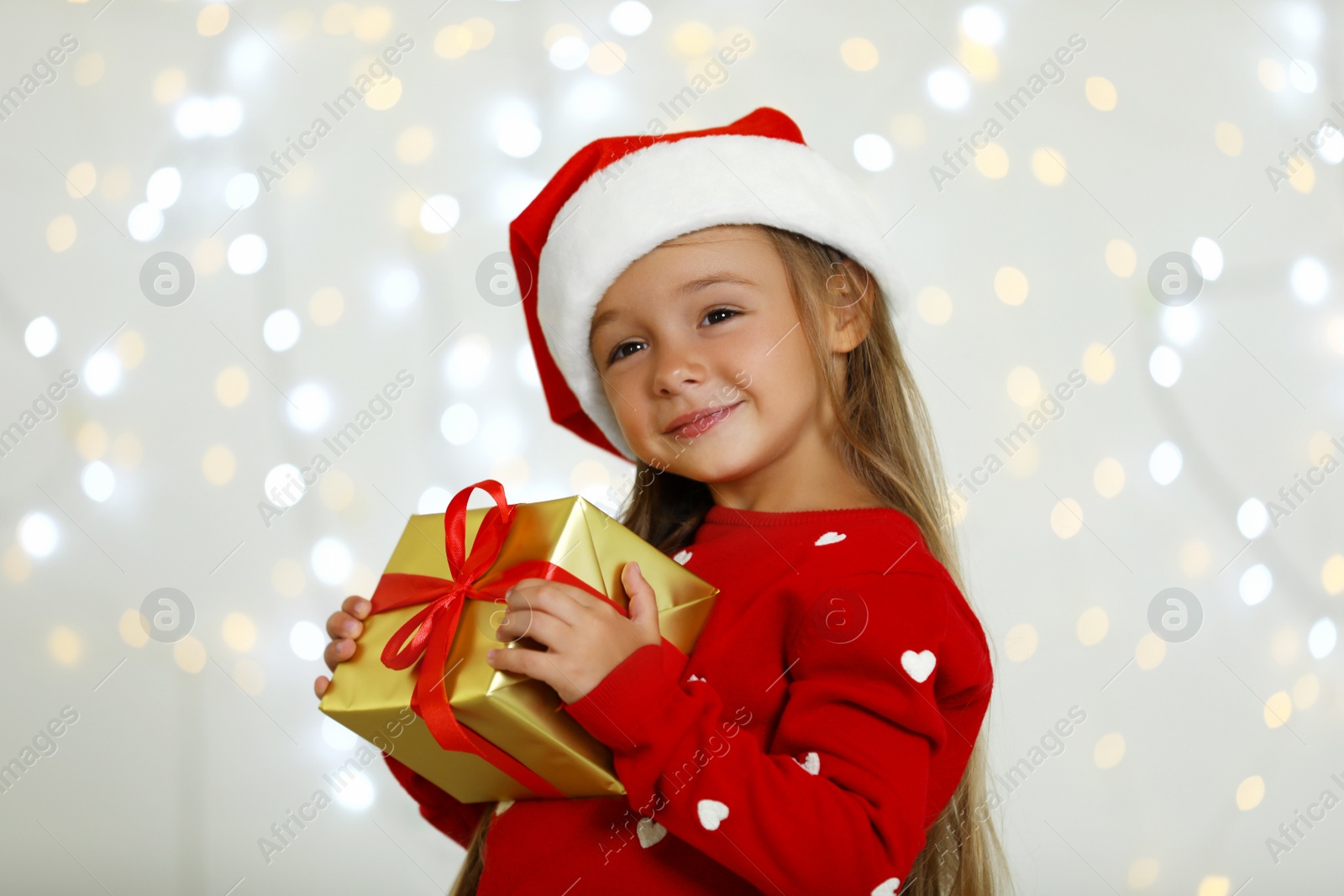 Photo of Happy little child in Santa hat with gift box against blurred festive lights. Christmas celebration