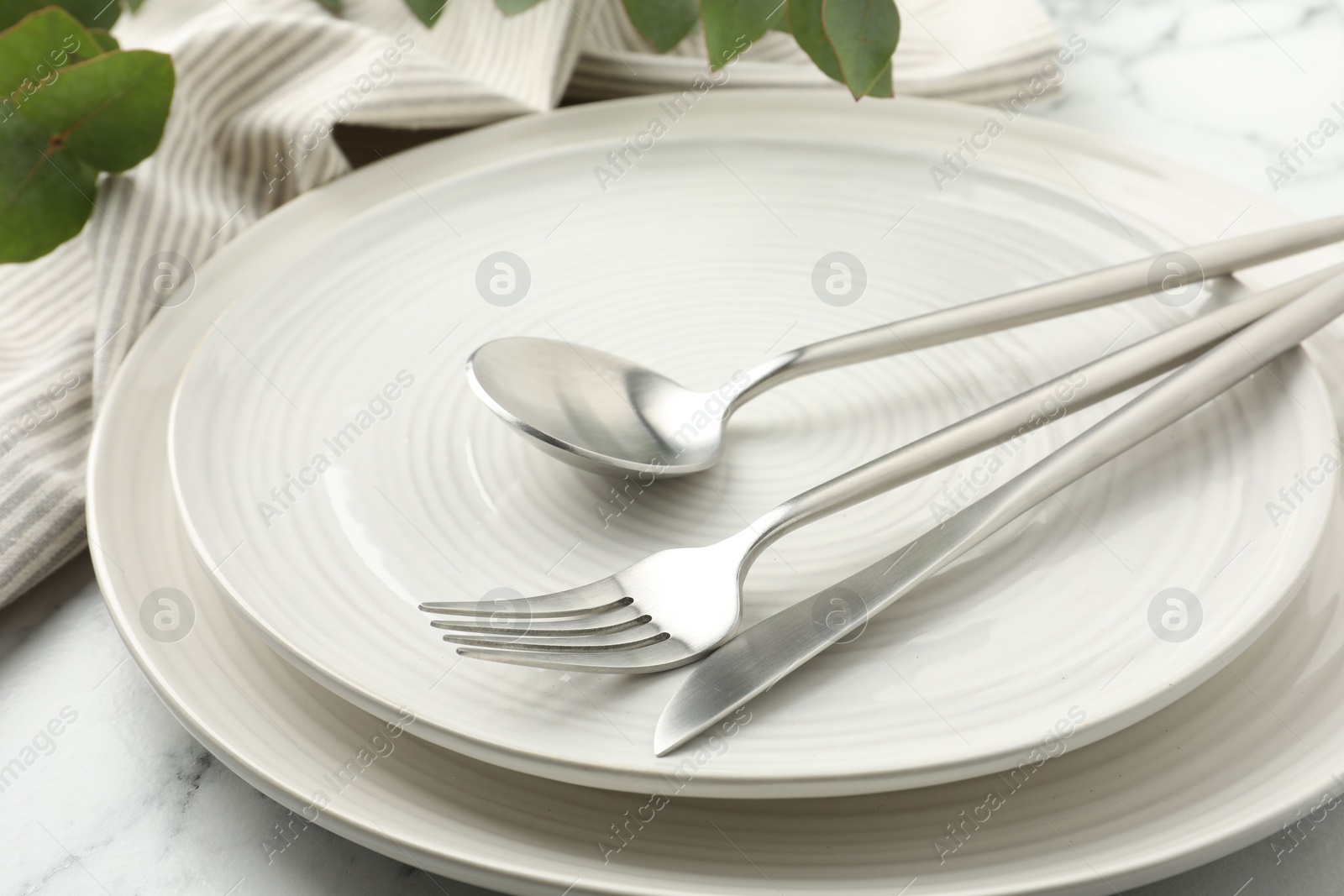 Photo of Stylish setting with cutlery, napkin, eucalyptus branches and plates on white marble table, closeup