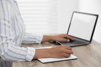 Woman writing notes while using laptop at wooden desk indoors, closeup