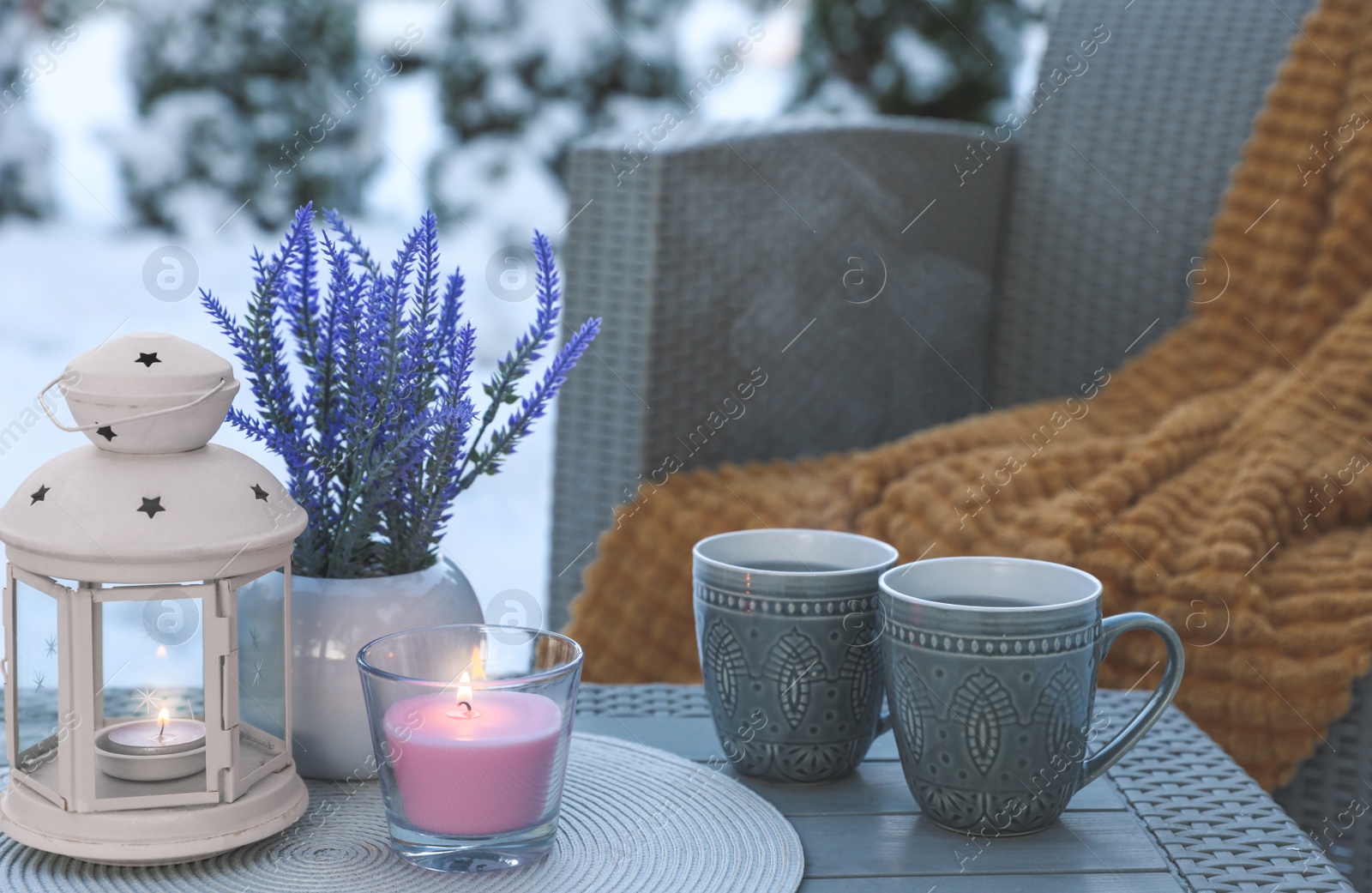 Photo of Burning candle, lantern, potted flowers and cups with hot drink on coffee table outdoors. Cosy winter