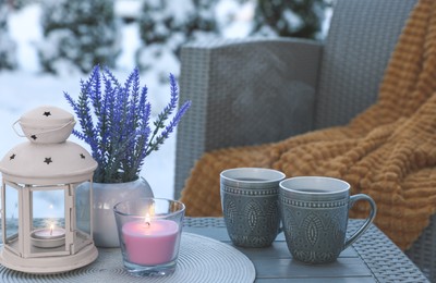 Burning candle, lantern, potted flowers and cups with hot drink on coffee table outdoors. Cosy winter