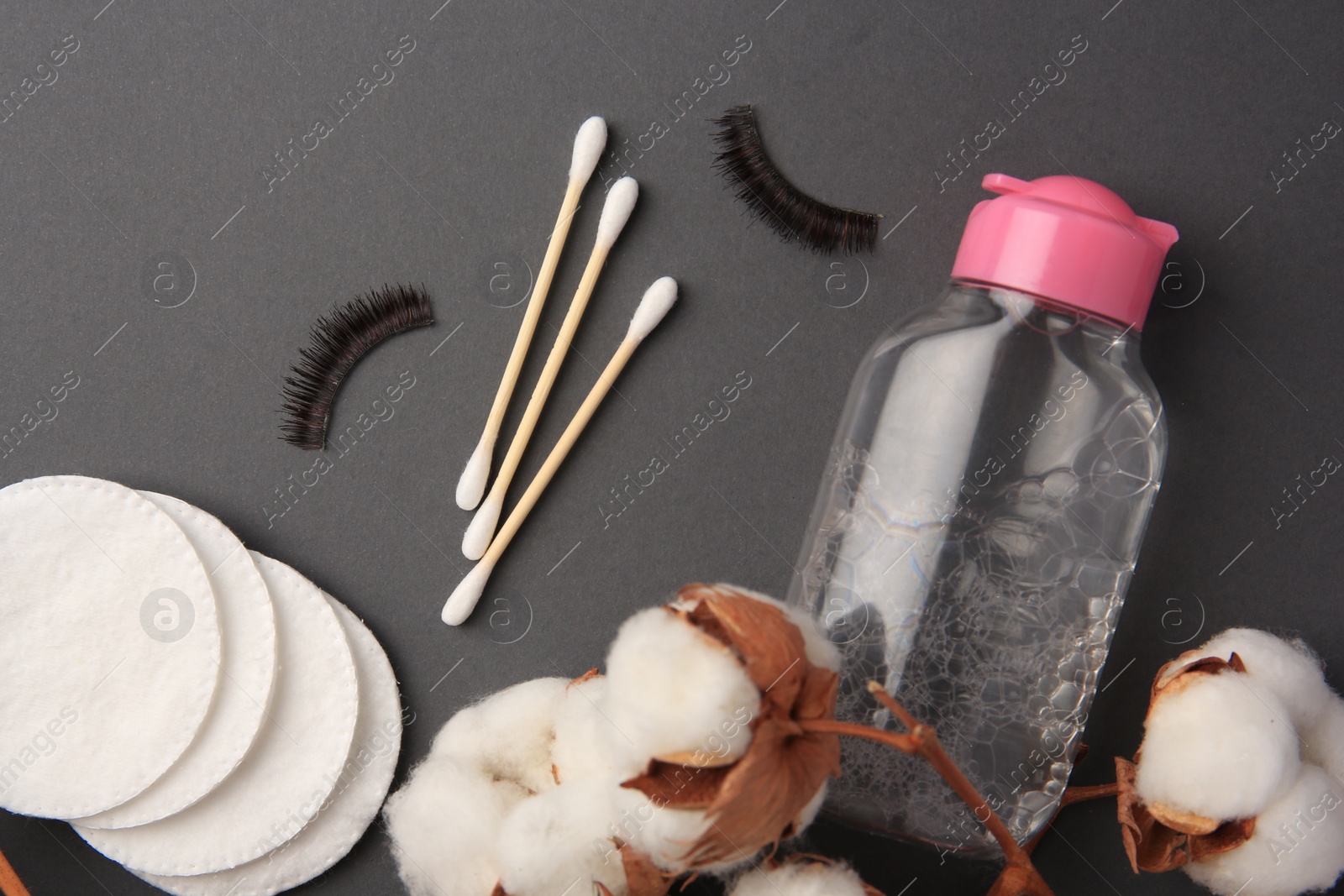 Photo of Bottle of makeup remover, cotton flowers, pads, swabs and false eyelashes on grey background, flat lay