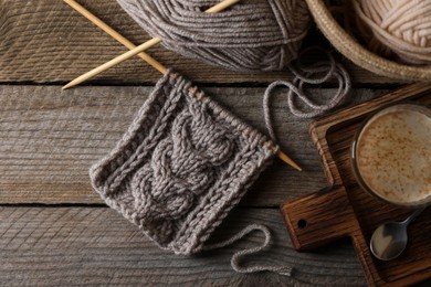 Soft grey woolen yarn, knitting, needles and glass of coffee on wooden table, flat lay