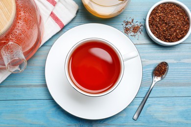 Photo of Freshly brewed rooibos tea and dry leaves on light blue wooden table, flat lay