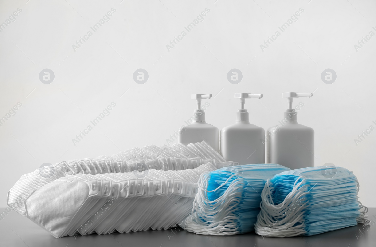 Photo of Hand sanitizers and respiratory masks on grey table against white background. Protective essentials during COVID-19 pandemic