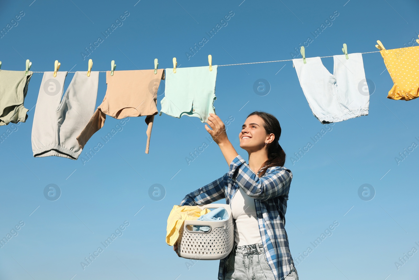 Photo of Smiling woman holding basket with baby clothes near washing line for drying against blue sky outdoors